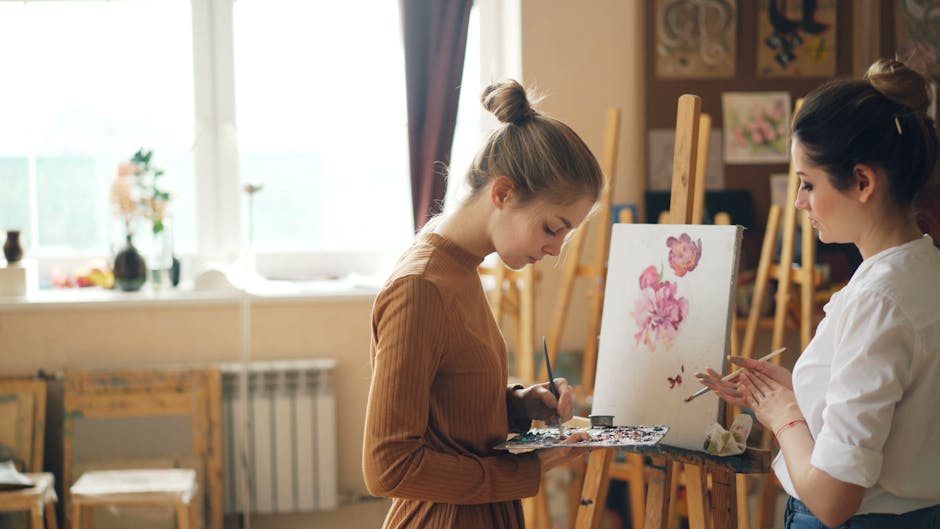 Two young women artists painting floral artwork on canvas in well-lit studio.