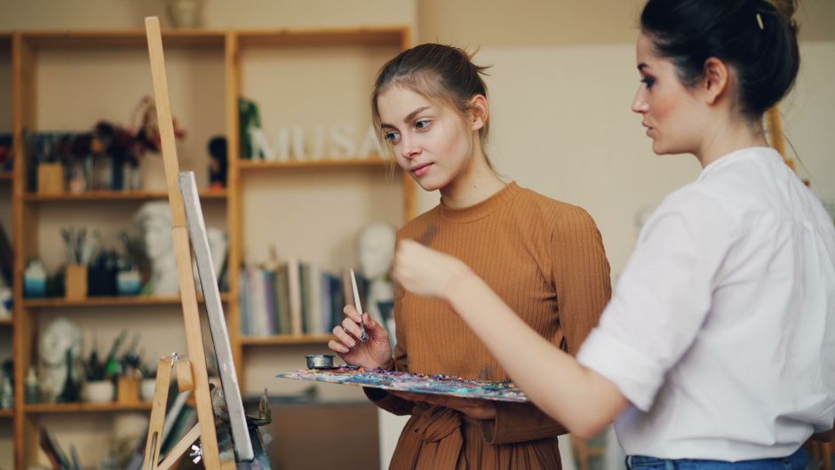Female art instructor teaching a young woman painting techniques in a cozy studio.